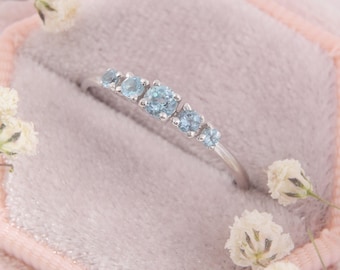 May include: A silver-toned ring featuring five round, light blue gemstones arranged in a row. The stones vary in size, with the largest stone positioned in the centre, flanked by two smaller stones on each side, all set on a thin, polished metal band.