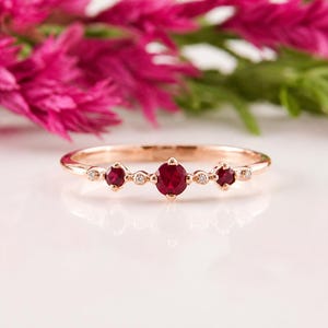 May include: A rose gold ring featuring three prominent round-cut red rubies set in a horizontal row, accented by four smaller clear round-cut stones interspersed between the rubies and at the outer edges of the band, creating a delicate design.