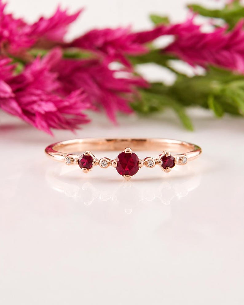 May include: A rose gold ring featuring three prominent round-cut red rubies set in a horizontal row, accented by four smaller clear round-cut stones interspersed between the rubies and at the outer edges of the band, creating a delicate design.