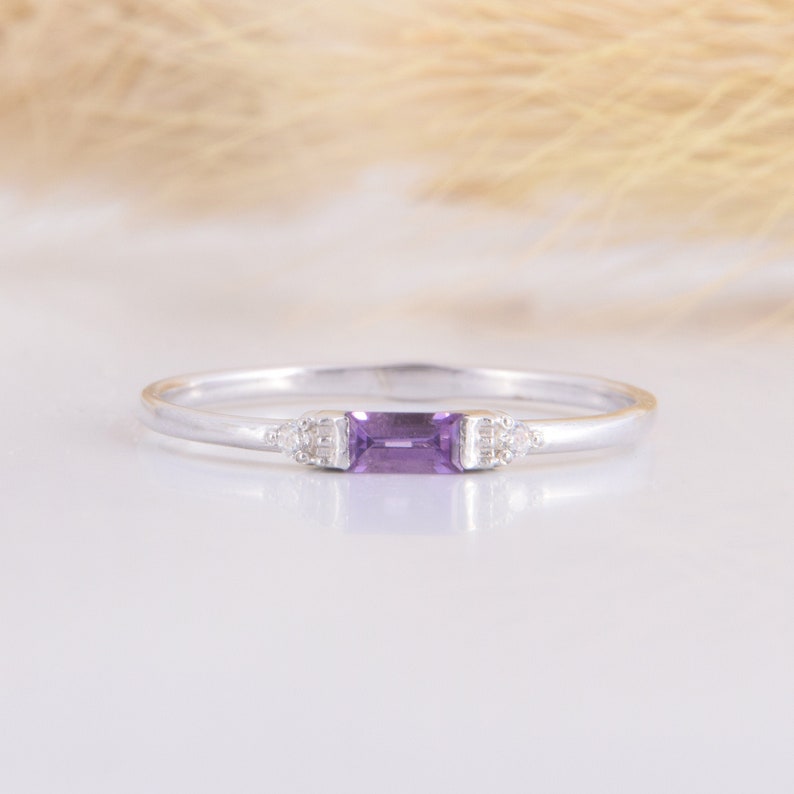 May include: A delicate silver ring with a rectangular purple amethyst gemstone and two smaller clear gemstones on either side.