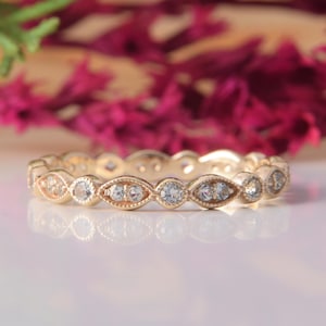 May include: A gold band ring featuring an alternating pattern of round and marquise-shaped settings, each containing a small clear gemstone. The metal has a beaded milgrain texture along the edges, creating a delicate and continuous design.