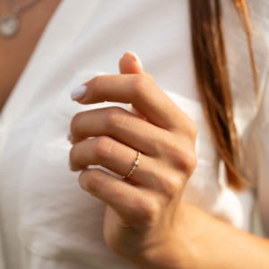 Pode incluir: Um delicado anel de ouro com uma pequena gema transparente engastada no centro. O anel &eacute; adornado com pequenas pedras brilhantes ao longo da banda.