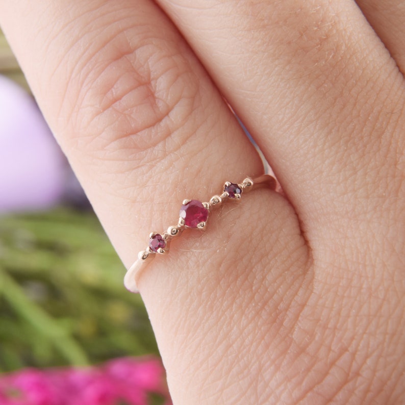 May include: A thin rose gold ring featuring three round, deep red rubies set in a row. The center ruby is slightly larger than the two smaller rubies on either side. The band is smooth and slender, showcasing a delicate and minimalist metal design.