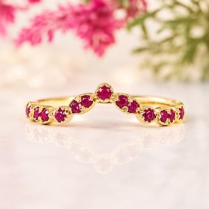 May include: A gold chevron-shaped ring featuring a series of small, round, vibrant pinkish-red gemstones set along the band. The ring has a polished metallic finish and a symmetrical V-shaped design that follows the curve of the band.
