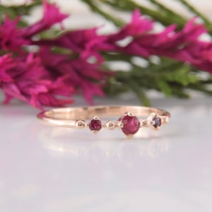 May include: A rose gold ring featuring a central round-cut red ruby flanked by two smaller red rubies. The band has a delicate, beaded texture along the setting, with the three stones arranged horizontally in a simple, polished metal design.