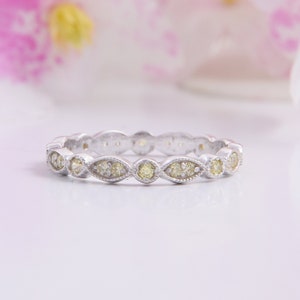 May include: A silver-toned band ring featuring a repeating pattern of marquise and circular settings. Each setting contains small, pale yellow stones. The metal band has a textured, beaded edge detail surrounding the stones in a continuous design.