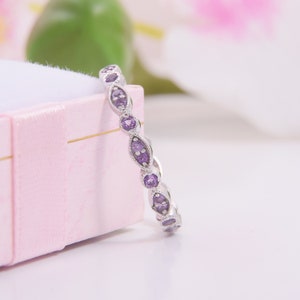 May include: A silver band ring featuring a series of alternating marquise and round-shaped purple gemstones set in a milgrain metal border. The ring is positioned upright against the side of a light pink rectangular jewellery box.