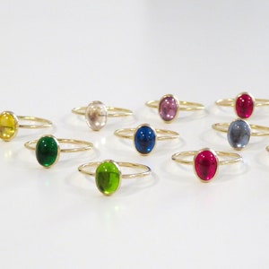 May include: A collection of 12 gold rings with oval-shaped gemstones in various colors, including green, blue, red, pink, yellow, and clear.