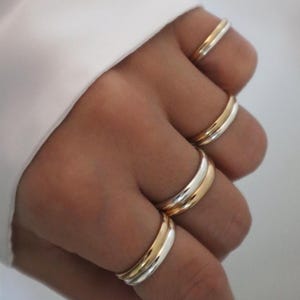 May include: A hand displaying multiple gold and silver rings. The rings are plain bands, some with a two-tone gold and silver design. The rings are worn on different fingers, creating a layered effect. The background is a white fabric.