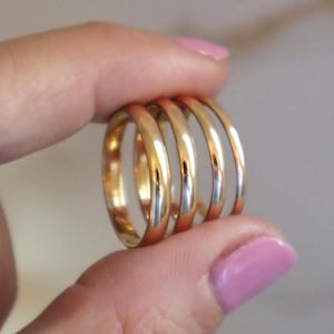 Può includere: Una collezione di cinque anelli d'oro, ciascuno con una fascia liscia e arrotondata. Gli anelli sono tenuti tra le dita, mostrando la loro superficie lucida e le diverse tonalità d'oro. Un gioiello classico.