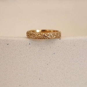 May include: A gold-colored ring with an intricate floral pattern. The ring is set against a neutral background, highlighting its detailed design. The band features a continuous, embossed pattern of leaves and vines.