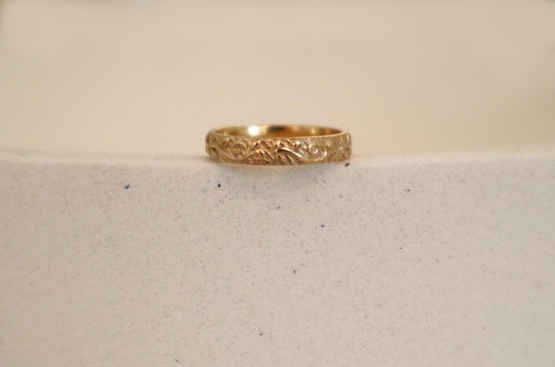 May include: A gold-colored ring with an intricate floral pattern. The ring is set against a neutral background, highlighting its detailed design. The band features a continuous, embossed pattern of leaves and vines.