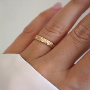 May include: A gold ring with an engraved floral pattern. The ring is worn on a finger, with a white fabric in the background. The ring's design features detailed leaves and swirls, showcasing a delicate and ornate style.