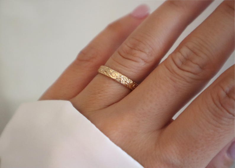 May include: A gold ring with an engraved floral pattern. The ring is worn on a finger, with a white fabric in the background. The ring's design features detailed leaves and swirls, showcasing a delicate and ornate style.