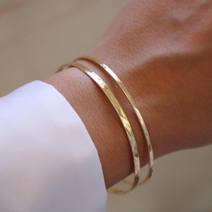 May include: Three gold-colored bangles are stacked on a wrist, with a white shirt sleeve visible in the foreground. The bangles have a hammered texture, reflecting light. The jewelry is simple and elegant.