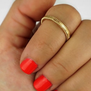 May include: A gold-toned ring with a textured, ribbed design. The ring is worn on the finger of a hand with red nail polish.