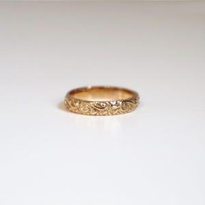 May include: A gold ring with an intricate floral pattern. The ring is a band style, with the design wrapping around the entire circumference. The ring is set against a white background, highlighting its details.