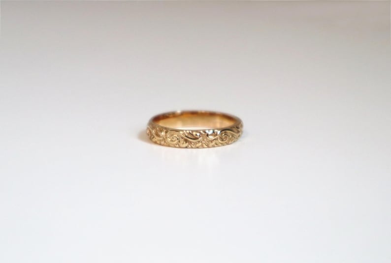 May include: A gold ring with an intricate floral pattern. The ring is a band style, with the design wrapping around the entire circumference. The ring is set against a white background, highlighting its details.