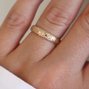 May include: A gold-colored ring with a detailed, textured pattern. The ring is worn on a finger, showcasing its intricate design. The ring's surface reflects light, highlighting the embossed details. The ring appears to be made of metal.