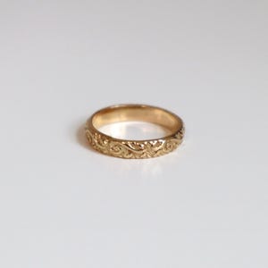 May include: A gold-colored ring with an intricate floral pattern. The ring is circular and appears to be made of metal. The design features swirling, leaf-like details around the band. The ring is set against a plain white background.