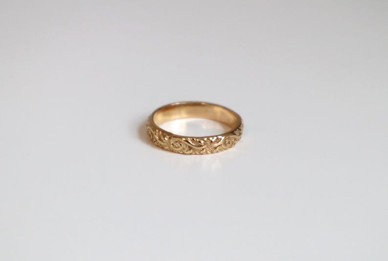 May include: A gold-colored ring with an intricate floral pattern. The ring is circular and appears to be made of metal. The design features swirling, leaf-like details around the band. The ring is set against a plain white background.
