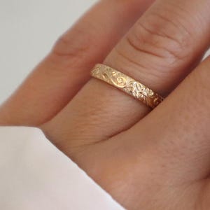 May include: A gold-colored ring with an engraved floral pattern. The ring is worn on a finger, showcasing the intricate details of the design. The ring's surface reflects light, highlighting the delicate carvings.
