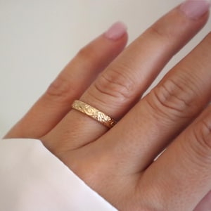May include: A gold ring with an engraved floral pattern. The ring is worn on a finger, showcasing its intricate design and warm color. The ring's details are highlighted against the skin and a white background.