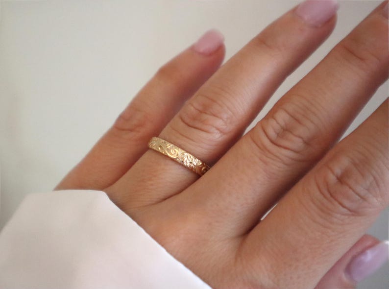 May include: A gold ring with an engraved floral pattern. The ring is worn on a finger, showcasing its intricate design and warm color. The ring's details are highlighted against the skin and a white background.
