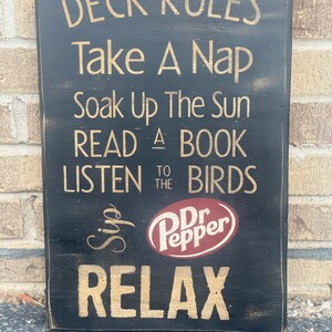 May include: Black wooden sign with gold lettering that reads "Deck Rules: Take a Nap, Soak Up The Sun, Read a Book, Listen to the Birds, Sip Dr Pepper, Relax".