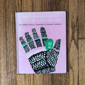 May include: A children's book titled "CATS HAVE KITTENS - DO GLOVES HAVE MITTENS?" features a pink cover with a hand illustration. The hand is black and white with green accents and a green mitten with a flower. The author is Cathleen Schurr.