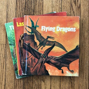May include: A stack of vintage children's books with dinosaur illustrations. The book on top is titled "Flying Dragons" with a vibrant orange cover featuring pterosaurs. Other books visible include "The Last" and a green-covered book with a jungle scene.