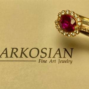 May include: A gold ring featuring a large oval ruby, encircled by small diamonds. A second gold band with small diamonds is next to it. The text "MARKOSIAN Fine Art Jewelry" is in the background.