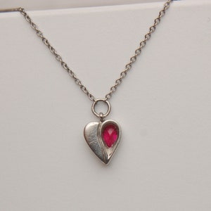 Może przedstawiać: Srebrny łańcuszek z naszyjnikiem w kształcie serca. Zawieszka ma czerwony kamień szlachetny w środku.