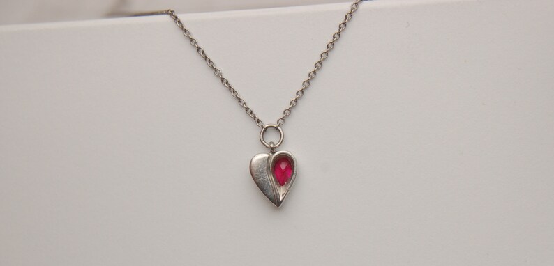 Może przedstawiać: Srebrny łańcuszek z naszyjnikiem w kształcie serca. Zawieszka ma czerwony kamień szlachetny w środku.