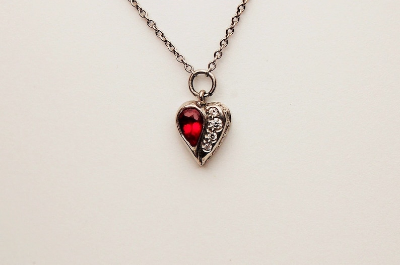 Może przedstawiać: Srebrny naszyjnik z serduszkowym wisiorem ozdobionym czerwonym kamieniem i przezroczystymi cyrkoniami.