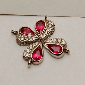 Może przedstawiać: Srebrny wisiorek w kształcie koniczyny czteroliściennej z czerwonymi kamieniami szlachetnymi i przezroczystymi cyrkoniami.