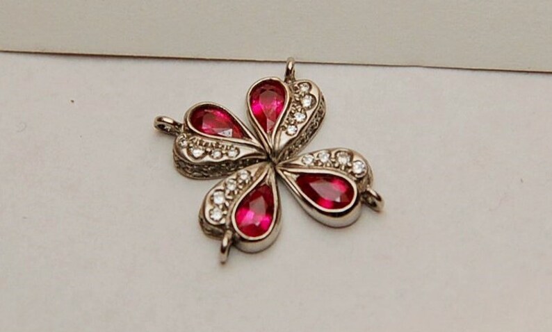 Może przedstawiać: Srebrny wisiorek w kształcie koniczyny czteroliściennej z czerwonymi kamieniami szlachetnymi i przezroczystymi cyrkoniami.