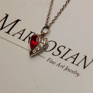 Może przedstawiać: Srebrny naszyjnik z sercem, ozdobiony czerwonym kamieniem szlachetnym i przezroczystymi kamieniami. Naszyjnik leży na białej kartce z tekstem "Marosian Fine Art Jewelry".