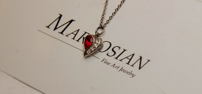 Może przedstawiać: Srebrny naszyjnik z sercem, ozdobiony czerwonym kamieniem szlachetnym i przezroczystymi kamieniami. Naszyjnik leży na białej kartce z tekstem "Marosian Fine Art Jewelry".