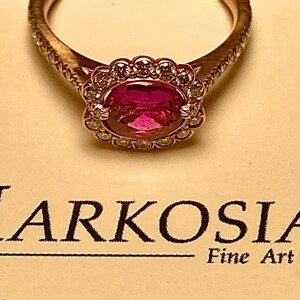 May include: Rose gold ring featuring a large oval ruby gemstone, encircled by a halo of smaller diamonds. The ring's band has a textured pattern. The text "Markosian Fine Art Jewelry" is visible below the ring.
