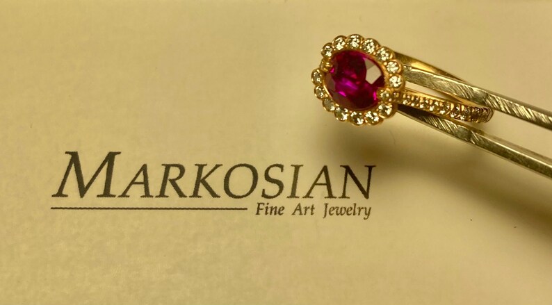 May include: A gold ring featuring a large oval ruby, encircled by small diamonds. A second gold band with small diamonds is next to it. The text "MARKOSIAN Fine Art Jewelry" is in the background.