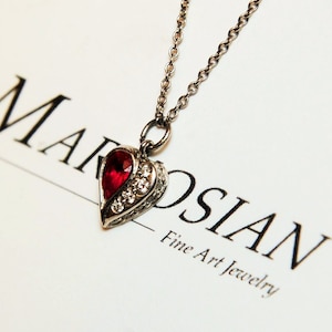 Może przedstawiać: Srebrny naszyjnik z sercowatym wisiorem ozdobionym czerwonym kamieniem szlachetnym i przezroczystymi cyrkoniami. Wisiorek znajduje się na białym tle z napisem "MAROSIAN Fine Art Jewelry".