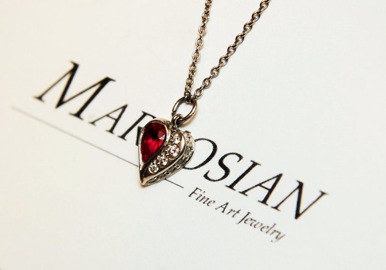 Może przedstawiać: Srebrny naszyjnik z sercowatym wisiorem ozdobionym czerwonym kamieniem szlachetnym i przezroczystymi cyrkoniami. Wisiorek znajduje się na białym tle z napisem "MAROSIAN Fine Art Jewelry".