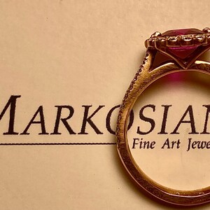 May include: A gold ring featuring a large, square-cut, pink gemstone, encircled by smaller stones. The ring rests on a cream surface with the text "Markosian Fine Art Jewelry".