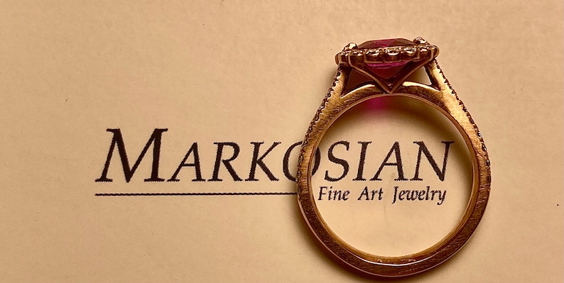 May include: A gold ring featuring a large, square-cut, pink gemstone, encircled by smaller stones. The ring rests on a cream surface with the text "Markosian Fine Art Jewelry".