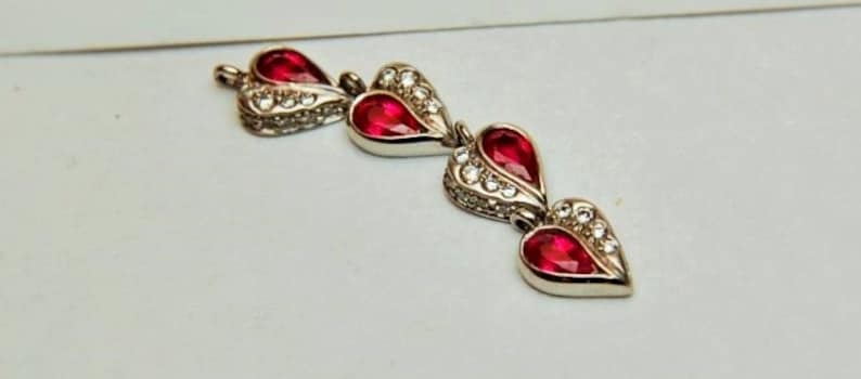 Może przedstawiać: Trzy srebrne zawieszki w kształcie serca z czerwonymi kamieniami szlachetnymi i przezroczystymi akcentami kryształowymi.
