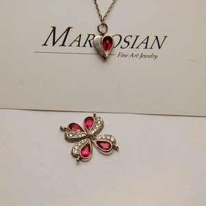 Może przedstawiać: Srebrny naszyjnik z czerwonym sercem i srebrnym kwiatem z czerwonymi kamieniami. Naszyjnik leży na białym tle z kartą z napisem "Marosian Fine Art Jewelry".