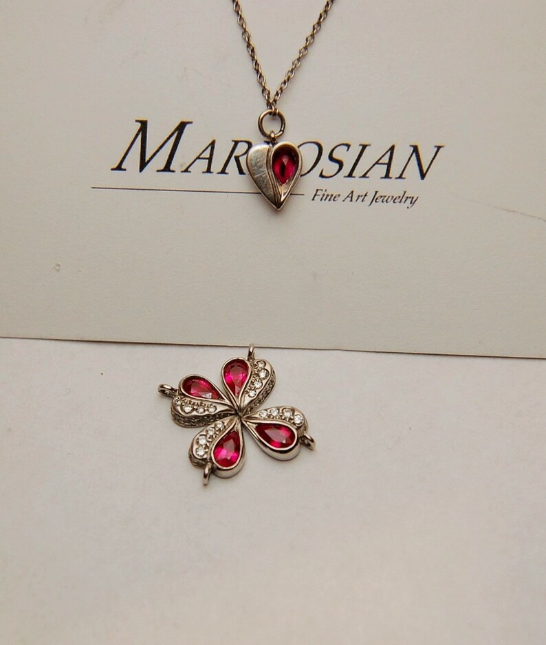 Może przedstawiać: Srebrny naszyjnik z czerwonym sercem i srebrnym kwiatem z czerwonymi kamieniami. Naszyjnik leży na białym tle z kartą z napisem "Marosian Fine Art Jewelry".