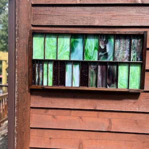 May include: A rectangular stained glass window with a dark brown wooden frame is mounted on a red-brown wooden wall. The glass panels feature shades of green, white, and brown. The window is divided into vertical sections.