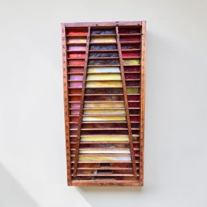 May include: A rectangular wooden printer's tray featuring a central trapezoidal section filled with horizontal slats. The compartments contain stained glass inserts in shades of red, orange, yellow, and brown, with numbers etched along the side edges.